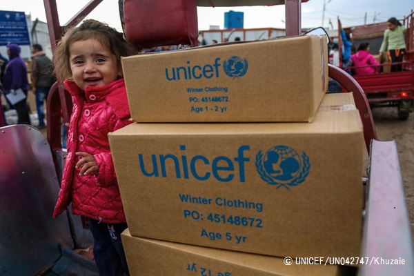 イラク・エルビルにある難民キャンプで支援物資の隣に笑顔で立つ、シリア人の女の子。（2016年12月1日撮影）