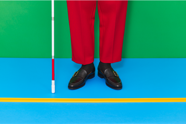 画像の説明：ココテープのビジュアル。背後の壁が緑、床は水色で黄色いココテープが横にまっすぐ引かれている。ココテープに両足を揃えて赤いズボン、黒いローファーを履き、白杖を持った人が立っている。