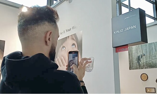 画像の説明：国際見本市にて男性が左手にKINJO JAPAN F0を曲げてもちスマートフォンで撮影している様子