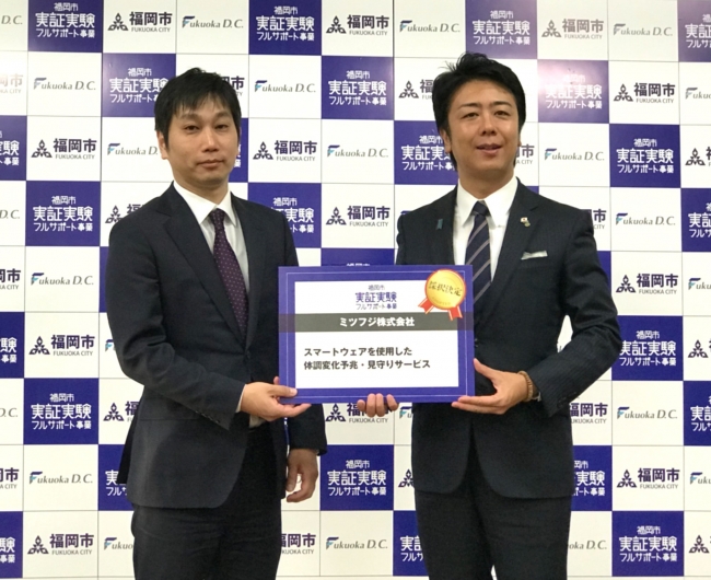 実証実験事業採択発表でパネルを渡す福岡市高島市長（右）とミツフジ三寺社長（左）