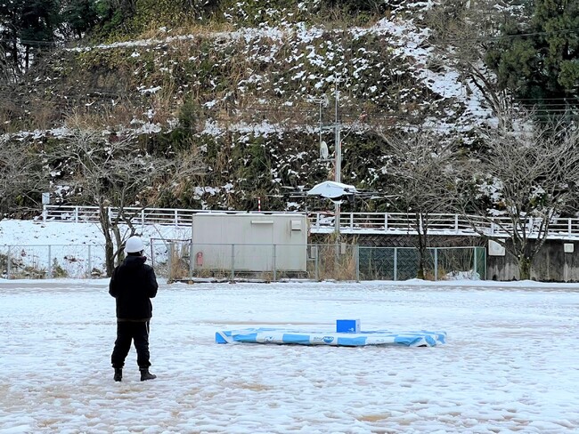 雪景色の中、児童生徒への荷物を届けて飛び立つドローン (松東みどり学園グラウンド) 雪景色の中、児童生徒への荷物を届けて飛び立つドローン (松東みどり学園グラウンド)