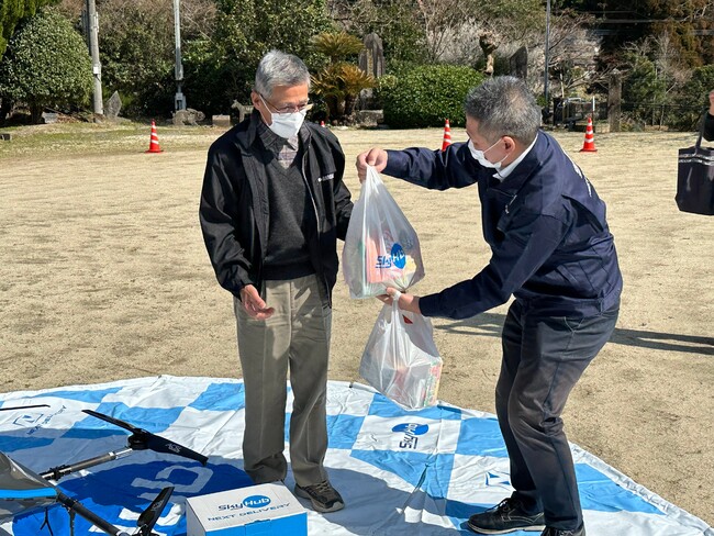 ドローン配送された健康食品セットを 受け取る地域住民モニター (高山区民会館にて) ドローン配送された健康食品セットを 受け取る地域住民モニター (高山区民会館にて)