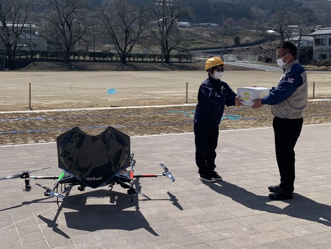 ドローン配送されたサイクリストのお土産が入った箱を陸送スタッフに手渡し (神川町多目的交流施設) ドローン配送されたサイクリストのお土産が入った箱を陸送スタッフに手渡し (神川町多目的交流施設)