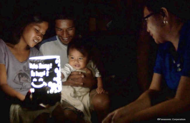 ソーラーランタンの明かりとシェードに描かれたパナソニック従業員のメッセージを見つめる家族