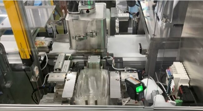 手前に積み上げた容器のふたをロボットアームが取り、惣菜の容器に押し付ける 手前に積み上げた容器のふたをロボットアームが取り、惣菜の容器に押し付ける