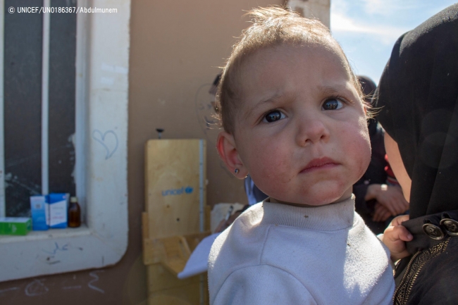 母親に抱えられ、集合型シェルターの外で待つ子ども。(2018年3月17日撮影) © UNICEF_UN0186367_Abdulmunem
