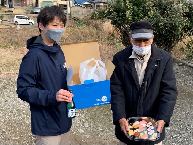 ドローン配送された「晩酌セット」のにぎり鮨が崩れていないことを見せる受け取った鈴木恒夫大沢区長 ドローン配送された「晩酌セット」のにぎり鮨が崩れていないことを見せる受け取った鈴木恒夫大沢区長